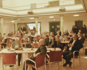 In einem Saal sitzen viele menschen an Tischen und schauen in die gleiche Richtung. Sie hören zu.