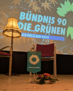 auf einer Bühne steht eine alte Stehlampe. Davor ein grüner Wimpel mit einer Sonnenblume darauf. Im Hintergrund auf einer Leinwand das Logo von BÜNDNIS 90/DIE GRÜNEN