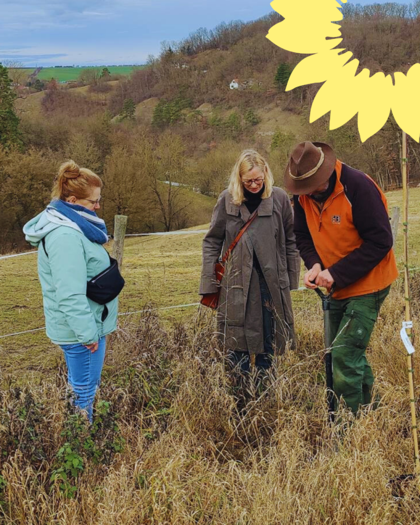 Auf einer herbstlichen Wiese stehen drei Menschen. Ein Mann mit Hut gräbt mit einem Spaten ein Loch. Daneben stehen zwei Frauen und unterhalten sich mit ihm