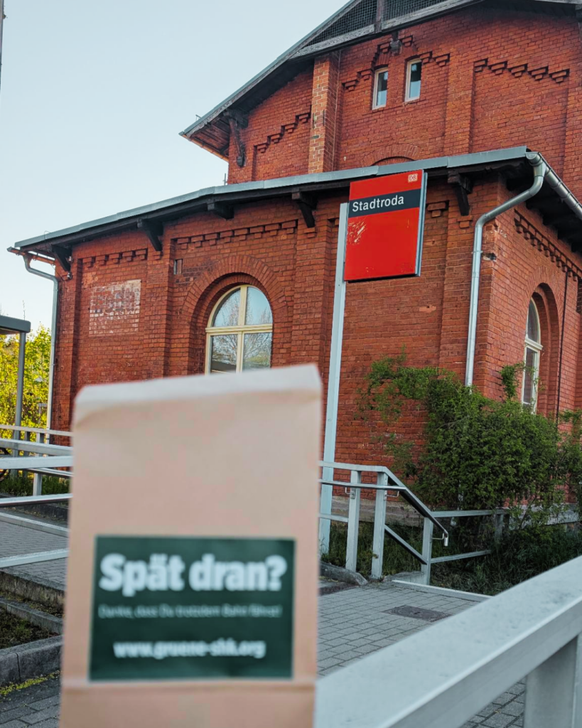 Foto vom Bahnhofsgebäude in Stadtroda. Da etwas unscharf ein Frühstücksbeutel mit dem Aufdruck: Spät dran? Danke, dass Du trotzdem Bahn fährst. www.gruene-shk.org